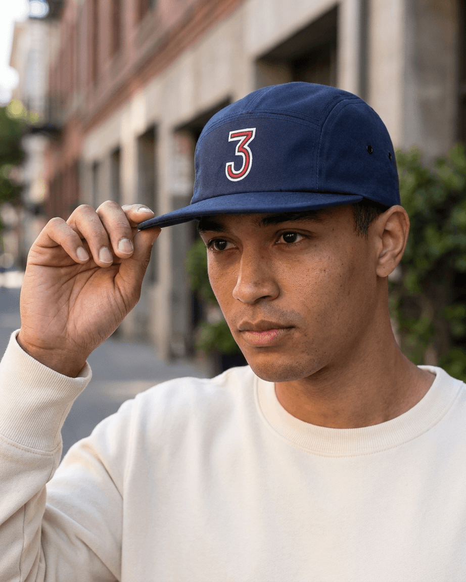 A man wearing a navy blue 5-panel baseball cap with a red and white number 3 on the front, holding the brim with his hand. He is wearing a cream-colored sweatshirt, with a blurred urban street background.