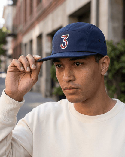 A man wearing a navy blue 5-panel baseball cap with a red and white number 3 on the front, holding the brim with his hand. He is wearing a cream-colored sweatshirt, with a blurred urban street background.