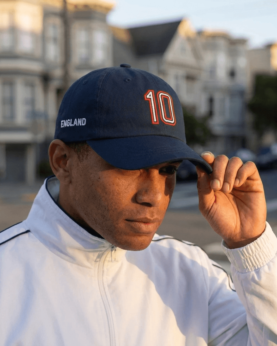A man wearing a navy blue England baseball cap with 'ENGLAND' and the number '10' in red and white, paired with a white zip-up jacket, set against a blurred background of residential buildings.