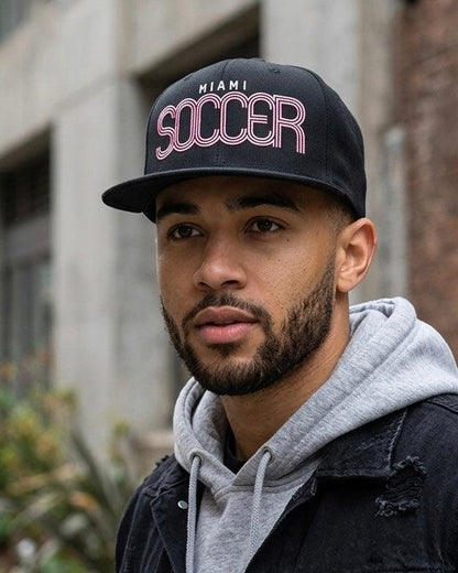 A man wearing a black snapback cap with 'MIAMI SOCCER' embroidered in pink and white on the front, paired with a gray hoodie and a dark distressed jacket, set against an urban background.