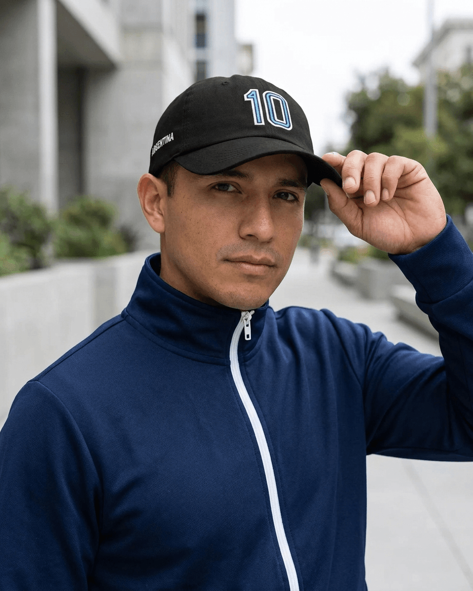 A man wearing a navy blue zip-up athletic jacket and a black baseball cap with the number 10 on it, standing outdoors in a city setting.