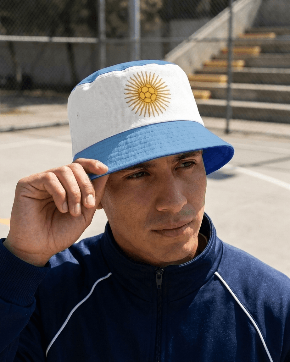A man wearing a white and light blue bucket hat with the Argentine sun emblem on the front, standing outdoors on a court with a fence and bleachers in the background.