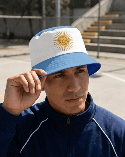 A man wearing a white and light blue bucket hat with the Argentine sun emblem on the front, standing outdoors on a court with a fence and bleachers in the background.