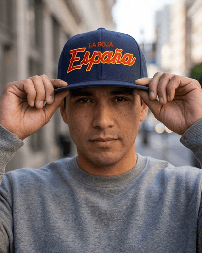 A man wearing a navy blue snapback cap with 'LA ROJA Española' embroidered in orange and white on the front, holding the brim with both hands in an outdoor urban setting.